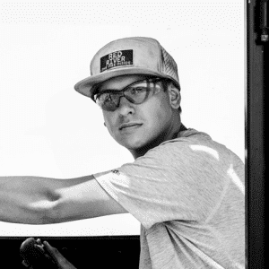 A person wearing a 'RED RIVER' cap and safety glasses, leaning on a vehicle or structure, dressed in a gray shirt.