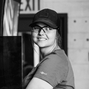 Woman in safety glasses and cap smiling while working indoors, wearing Force gear with EXIT sign visible in the background.