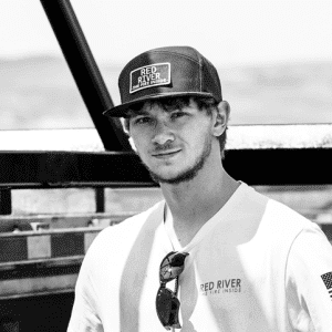 Man in Red River branded hat and shirt standing outdoors by metal structure, with sunglasses clipped to his collar.