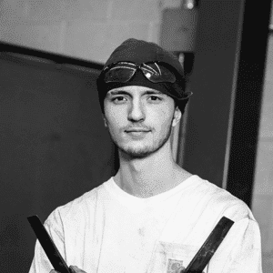 Young male welder wearing a beanie and safety goggles, holding metal bars while standing in a workshop.