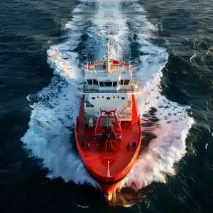 Aerial view of a large red ship with white and blue details sailing on the ocean, leaving a wake behind it.