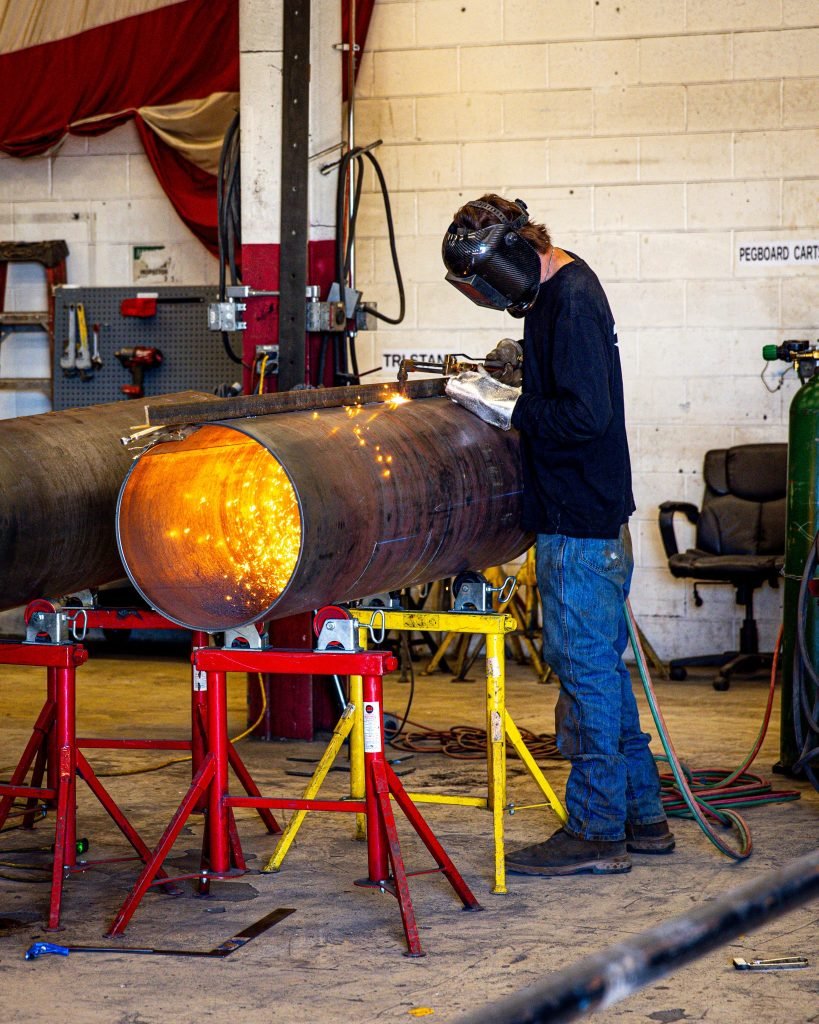 A welder in protective gear works on a large metal pipe, with bright sparks flying inside a workshop setting.