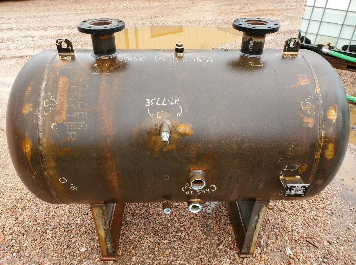 Horizontal cylindrical steel pressure vessel with multiple pipe nozzles, lifting lugs, and visible weld marks on gravel.