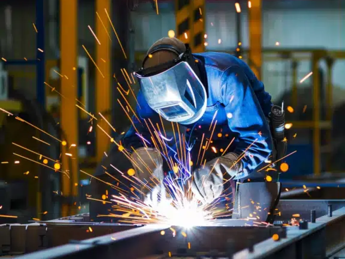 Industrial welder in protective gear working on metal structure, with bright sparks flying in a busy factory environment.