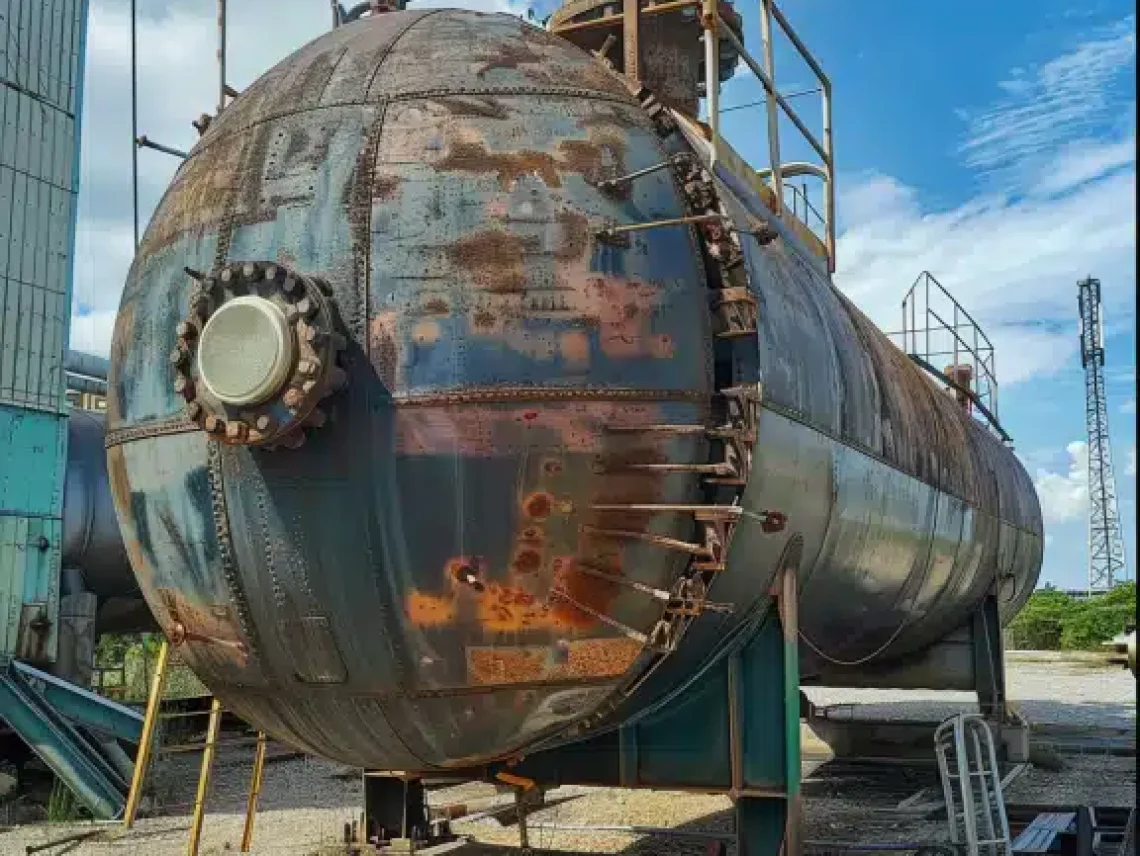 A large, rusted industrial pressure vessel or tank, surrounded by scaffolding, located outdoors with a cloudy sky.