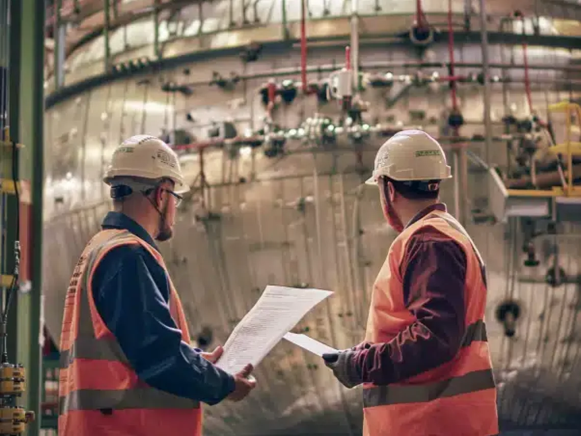 Two workers in safety vests and helmets review documents near a large industrial vessel in a factory setting.