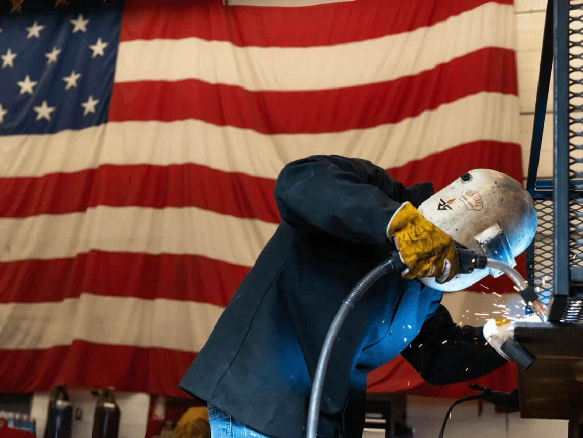 Red River welder in protective gear working with sparks flying in front of a large American flag, representing Red River’s skilled craftsmanship, pressure vessel fabrication, and American industrial manufacturing values.